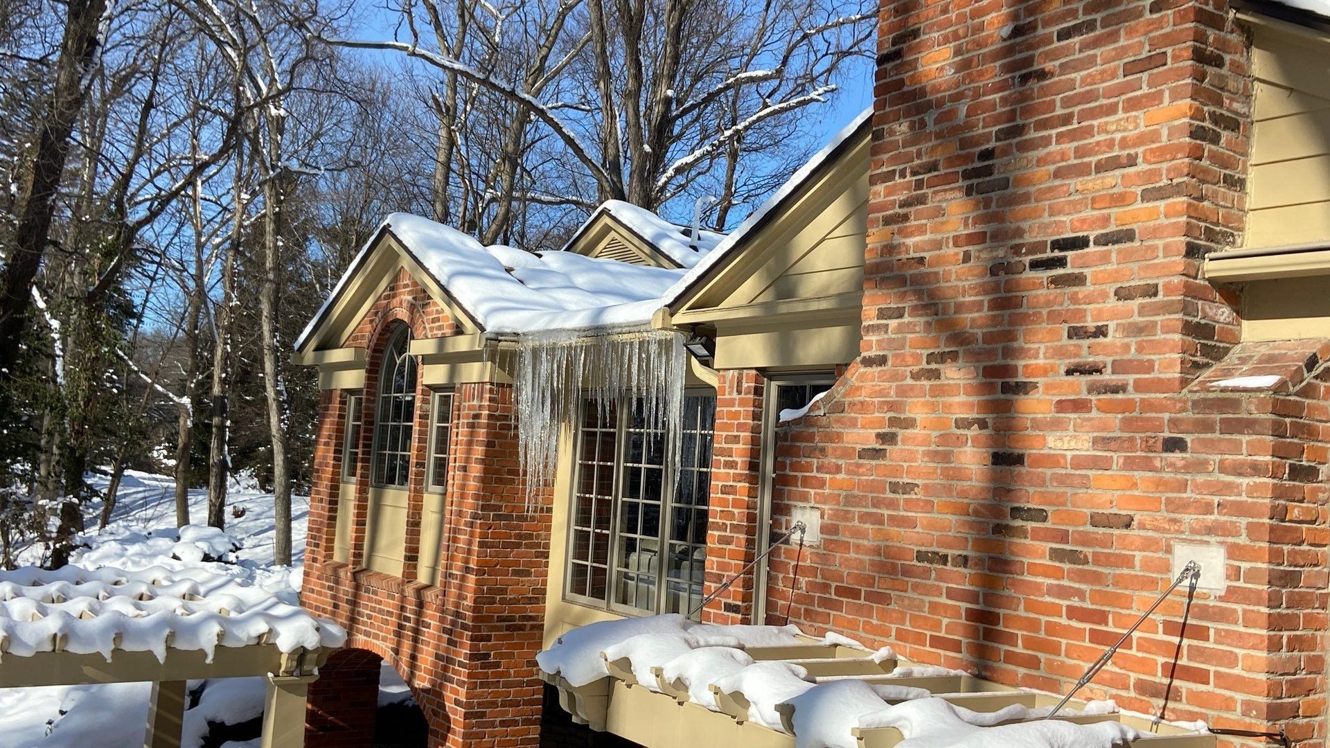 Brick house with snow on the roof and ground, seen in a winter wooded yard.