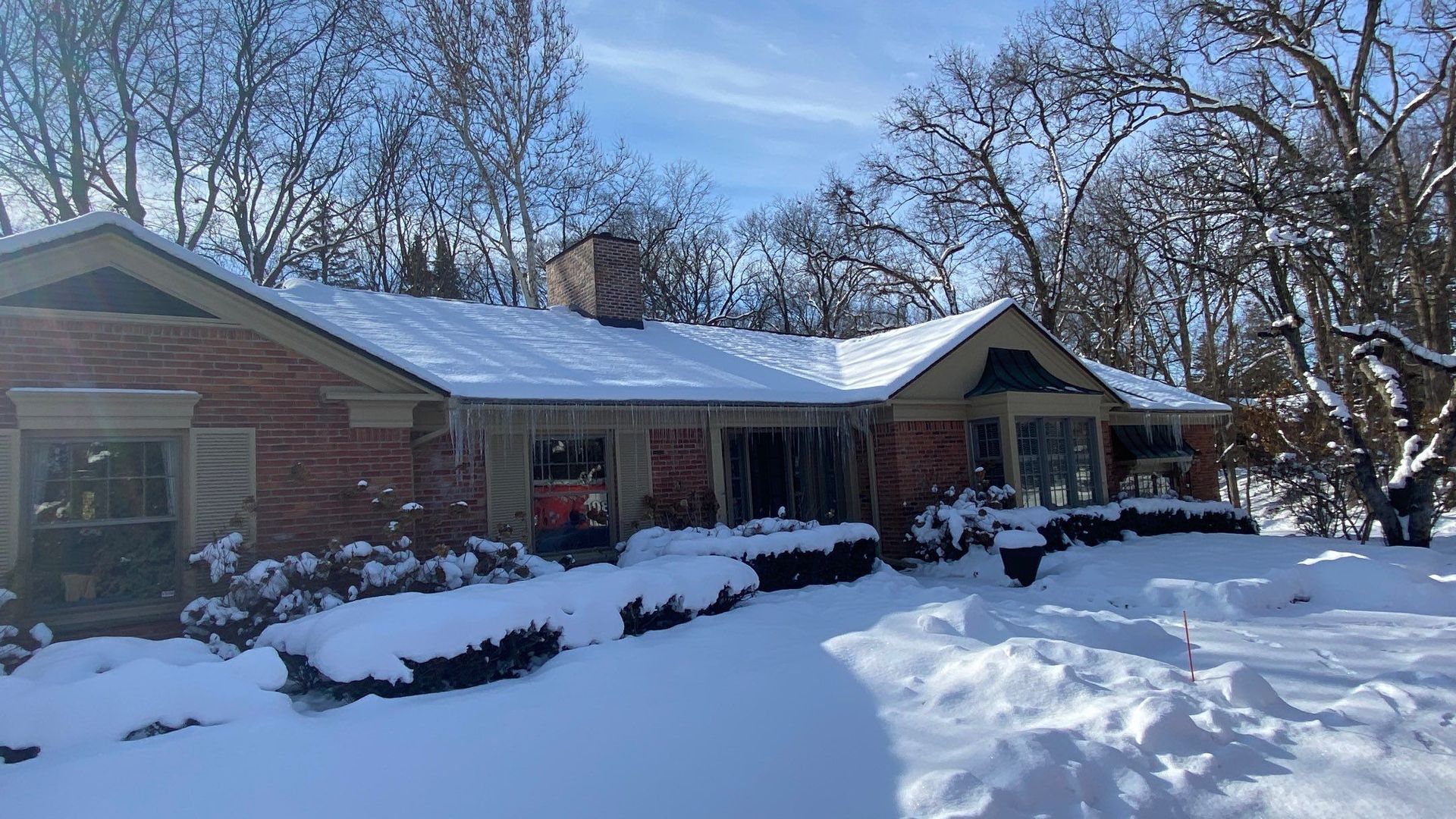 Brick house covered in deep snow under a clear blue sky, with bare trees in the background