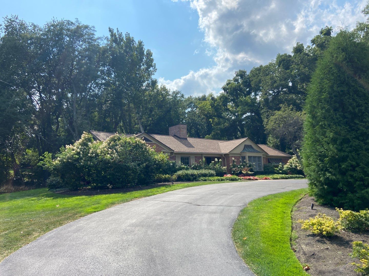 Curved driveway leading to a house with trees, shrubs, and a partly cloudy sky
