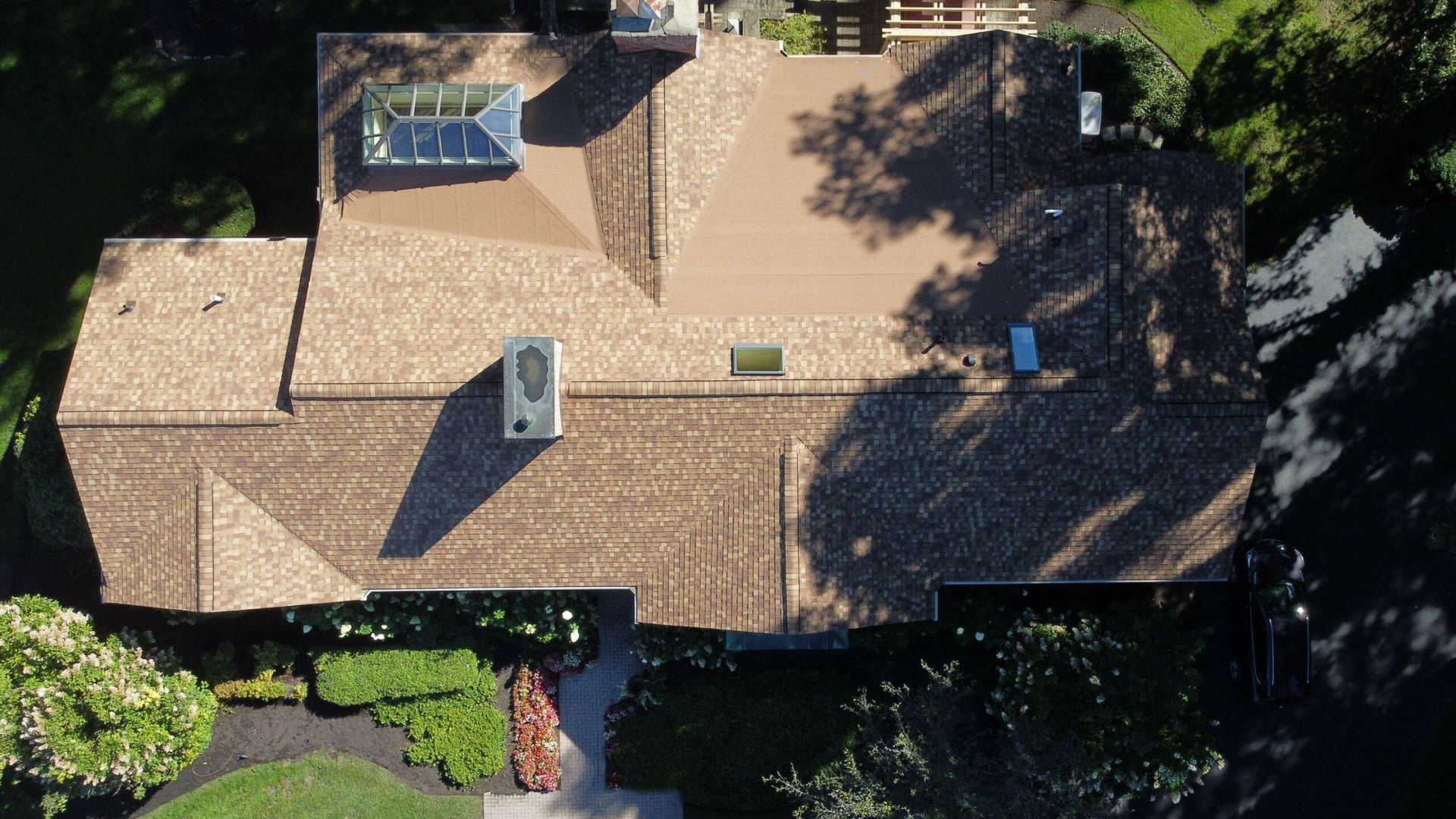 Aerial view of a tan shingled house roof with skylights, chimneys, and surrounding trees