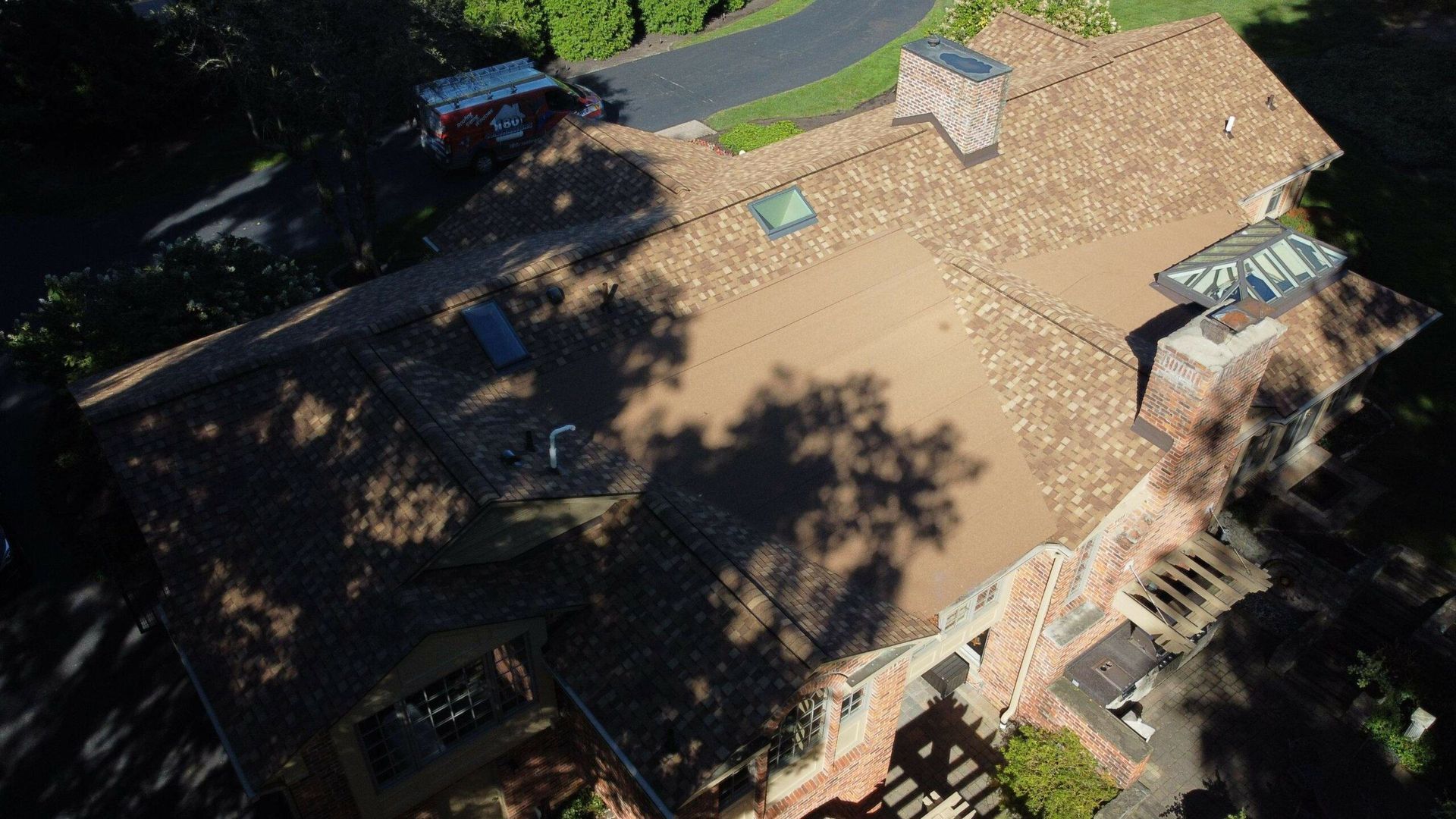 Aerial view of a tan-roofed house with tree shadows and a street visible nearby.