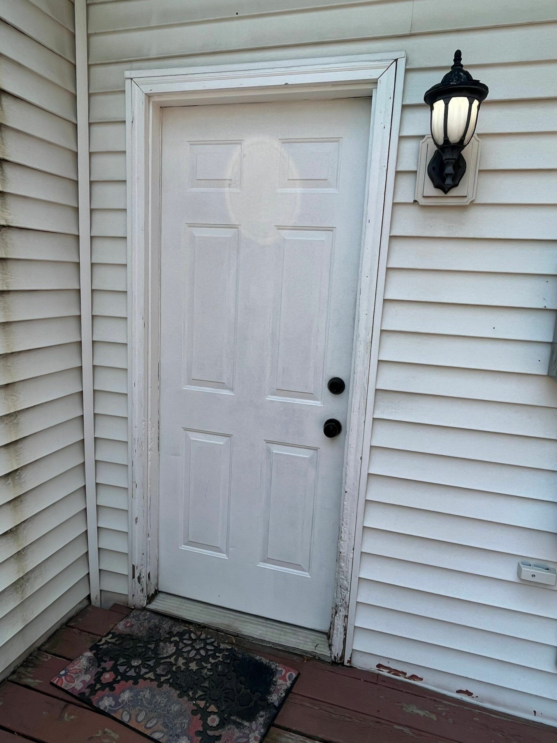 White front door with black lantern light on beige siding and a small welcome mat by the entrance