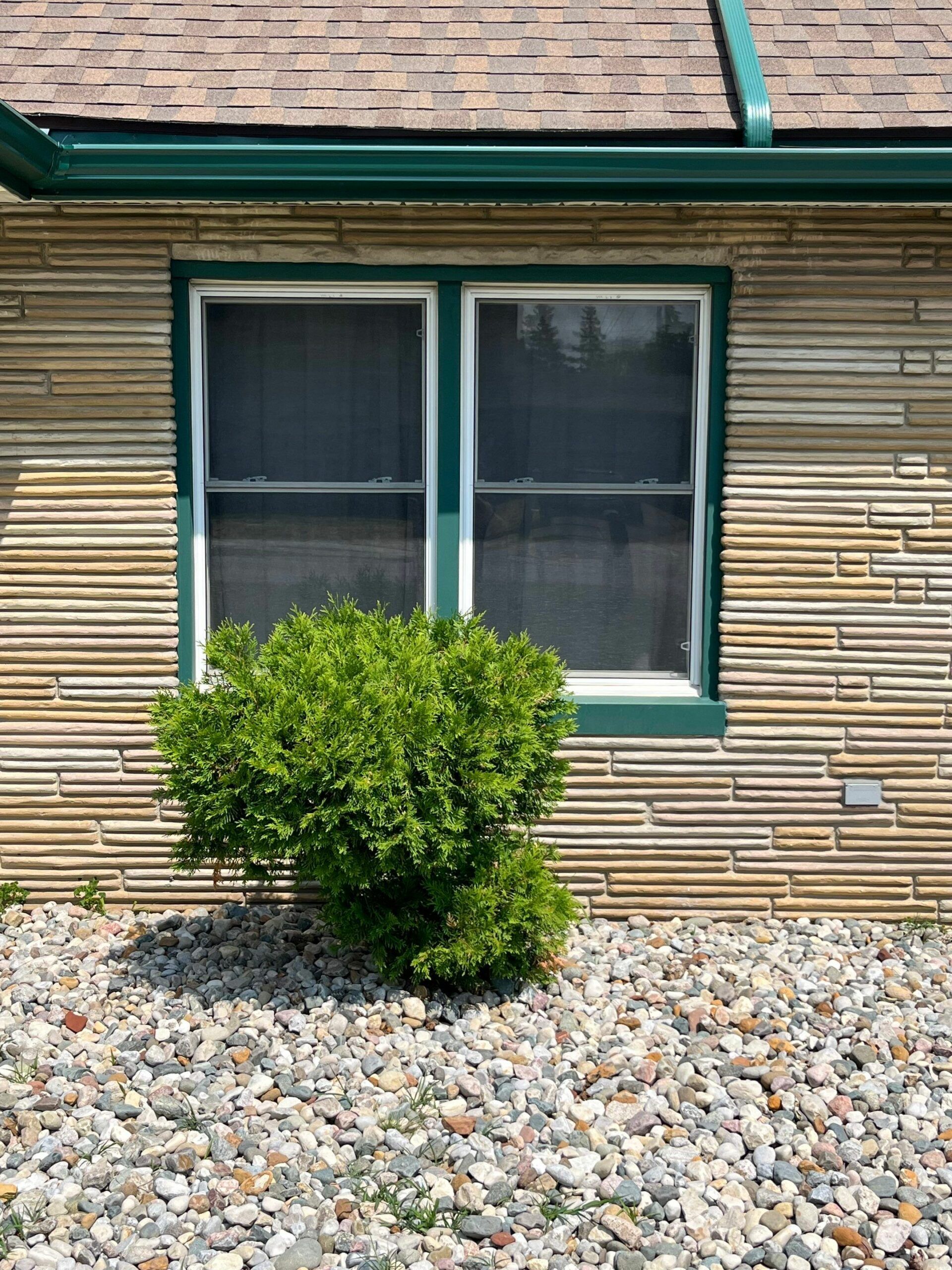 Exterior wall with a green-trimmed window above a small bush and rock landscaping.