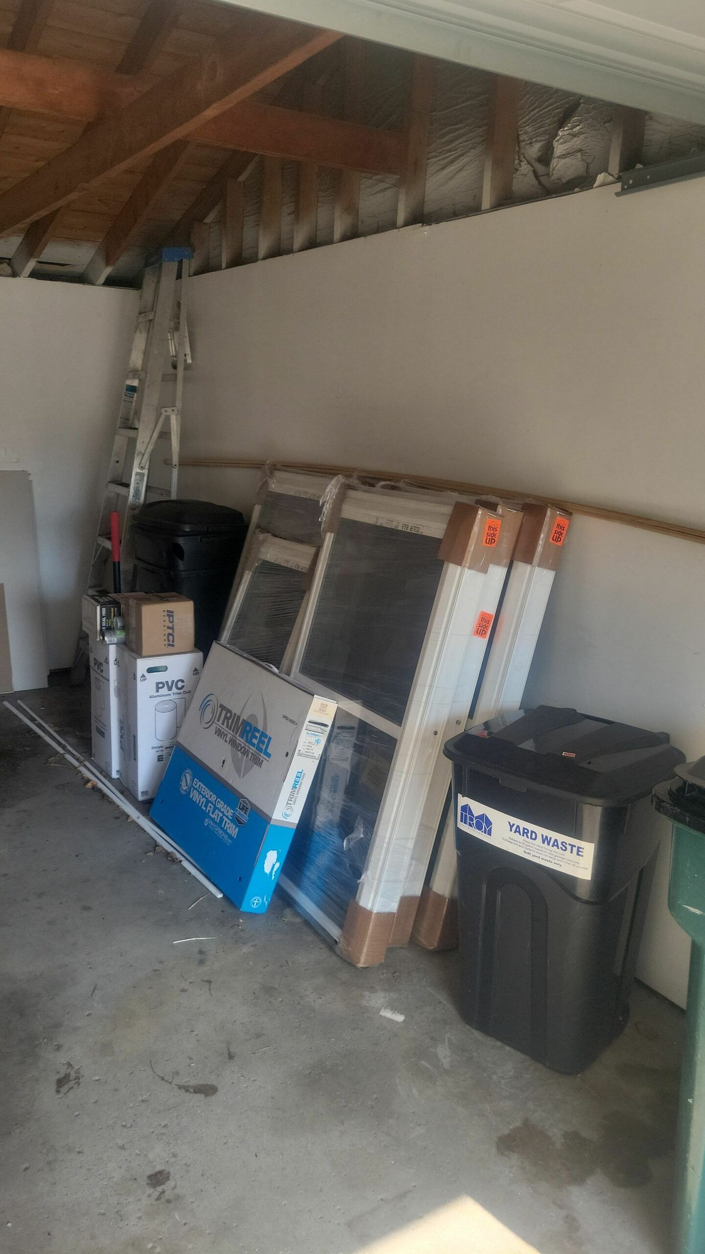 Garage with stacked construction materials, a ladder, and trash bins along the wall.