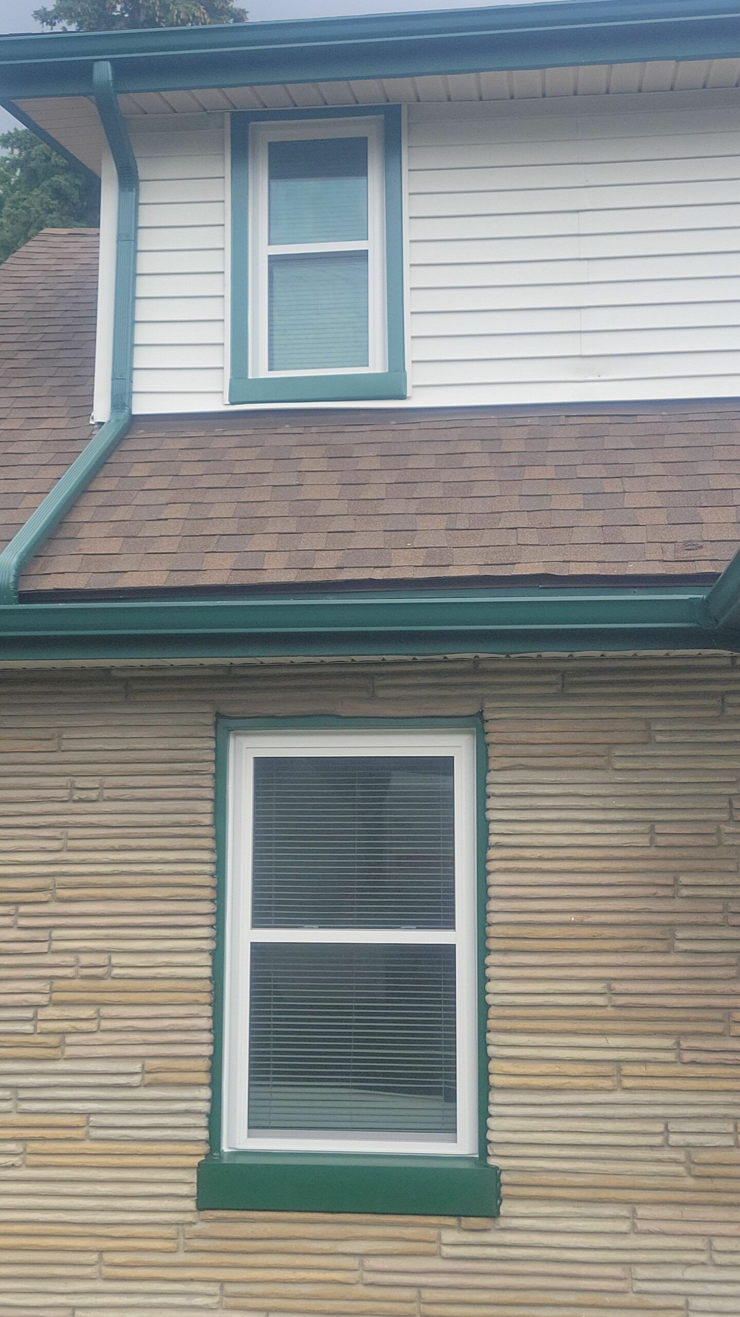Two-story house exterior with white siding, tan brick, green trim, and two windows.