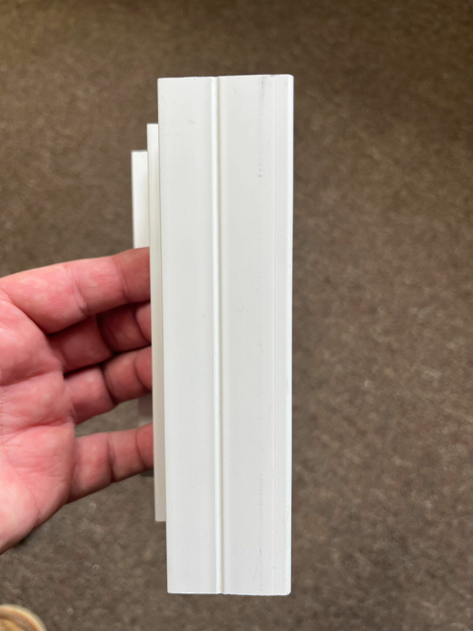 Hand holding a white vertical plastic trim piece against a brown carpet background