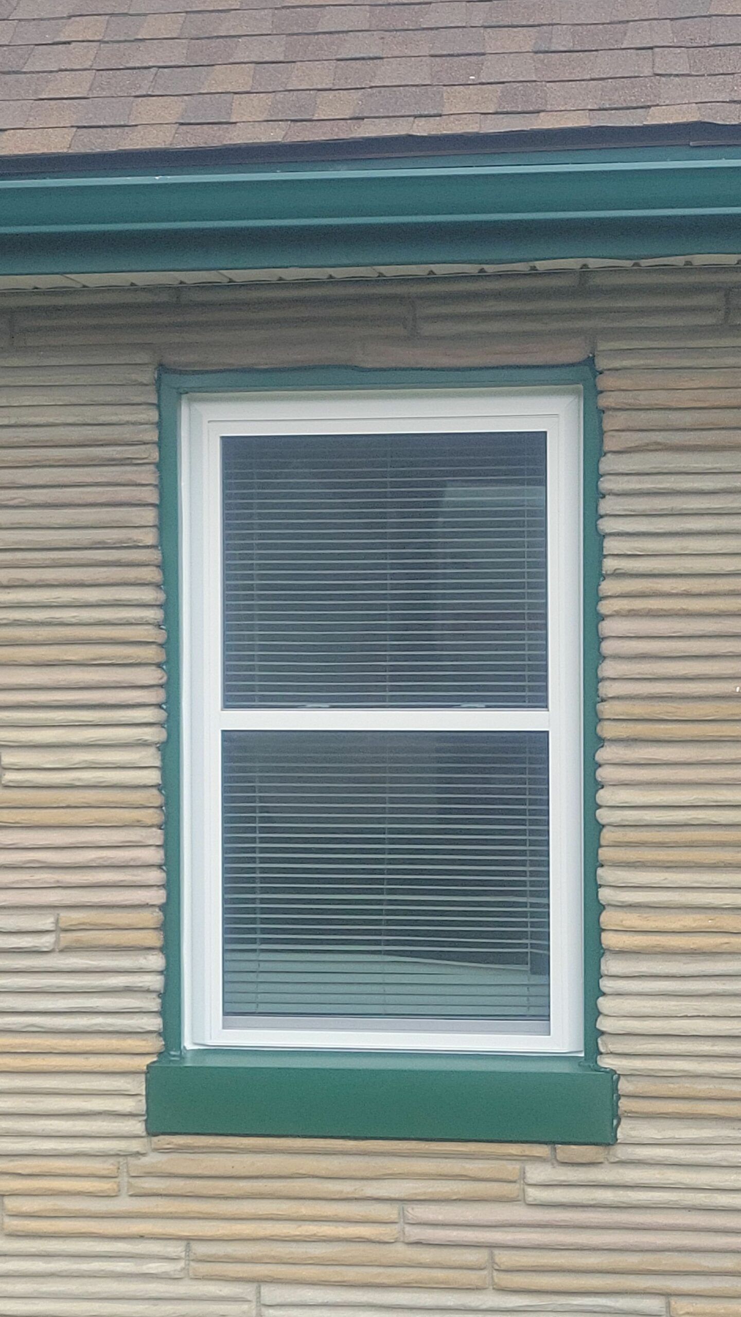 Single window with white blinds and green trim on a beige house exterior