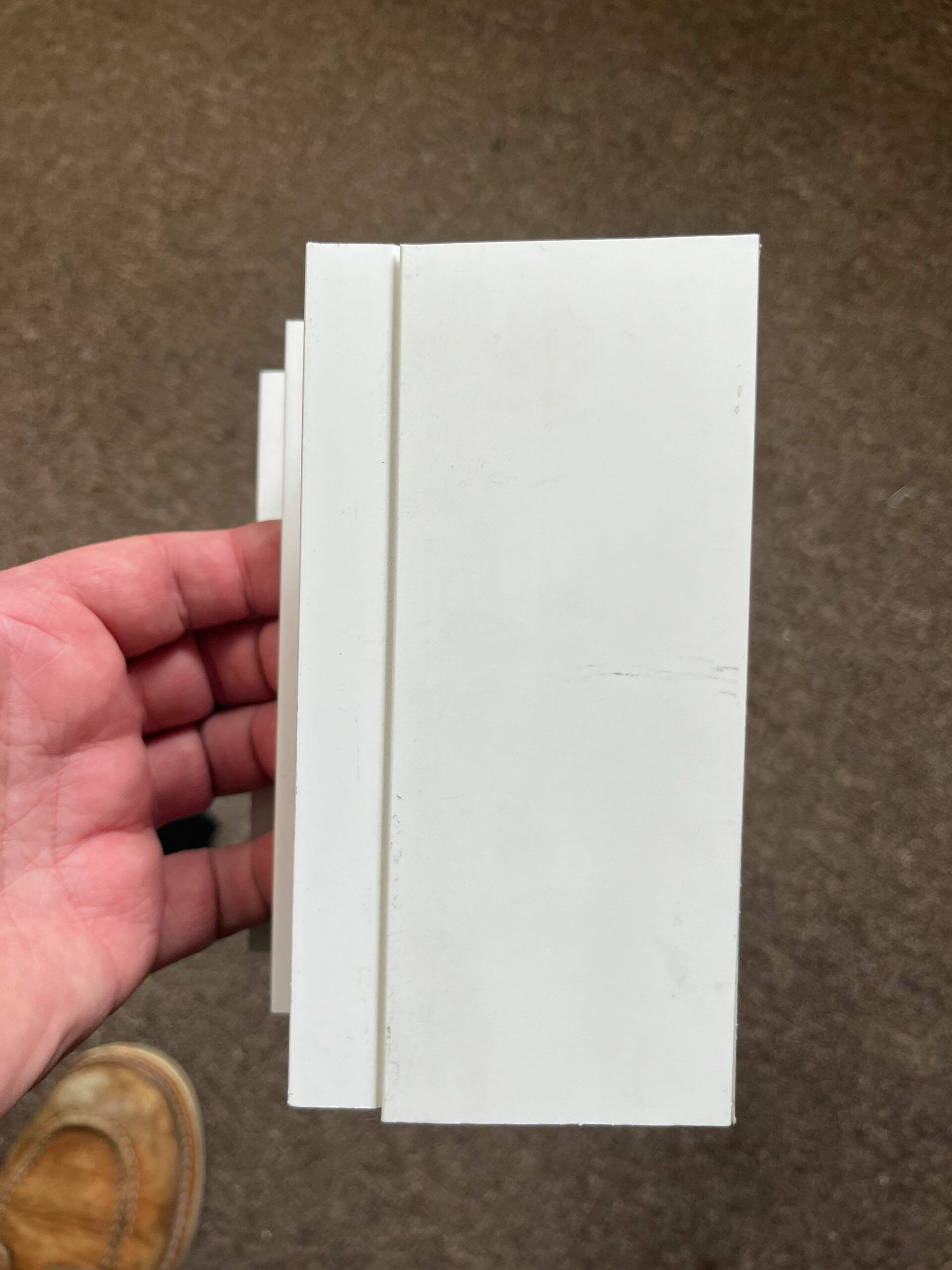 Hand holding two white rectangular panels on a carpeted floor, with a tan shoe visible nearby