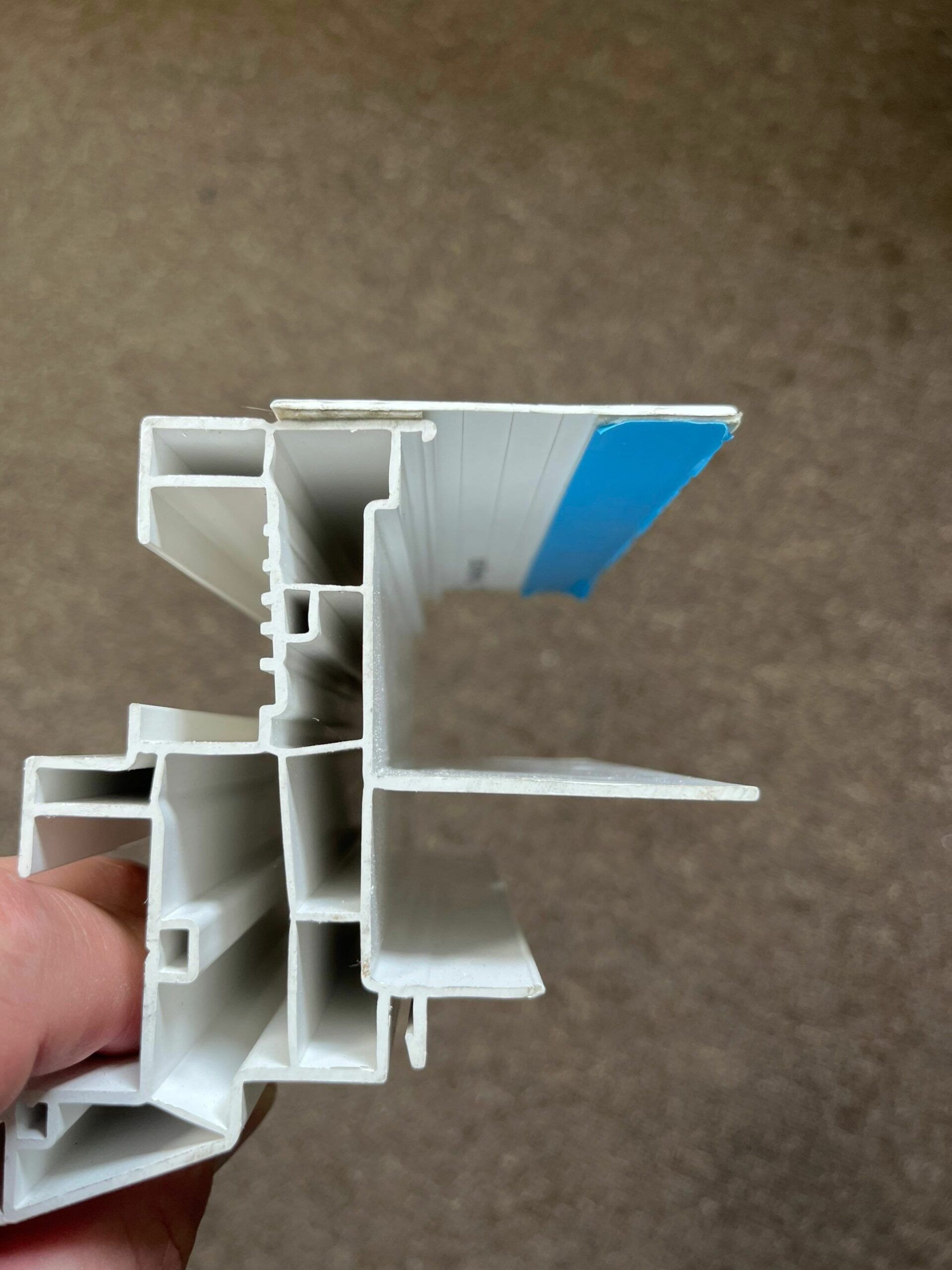 Cross-section of a white plastic profile with blue tape, held by a hand against a beige background