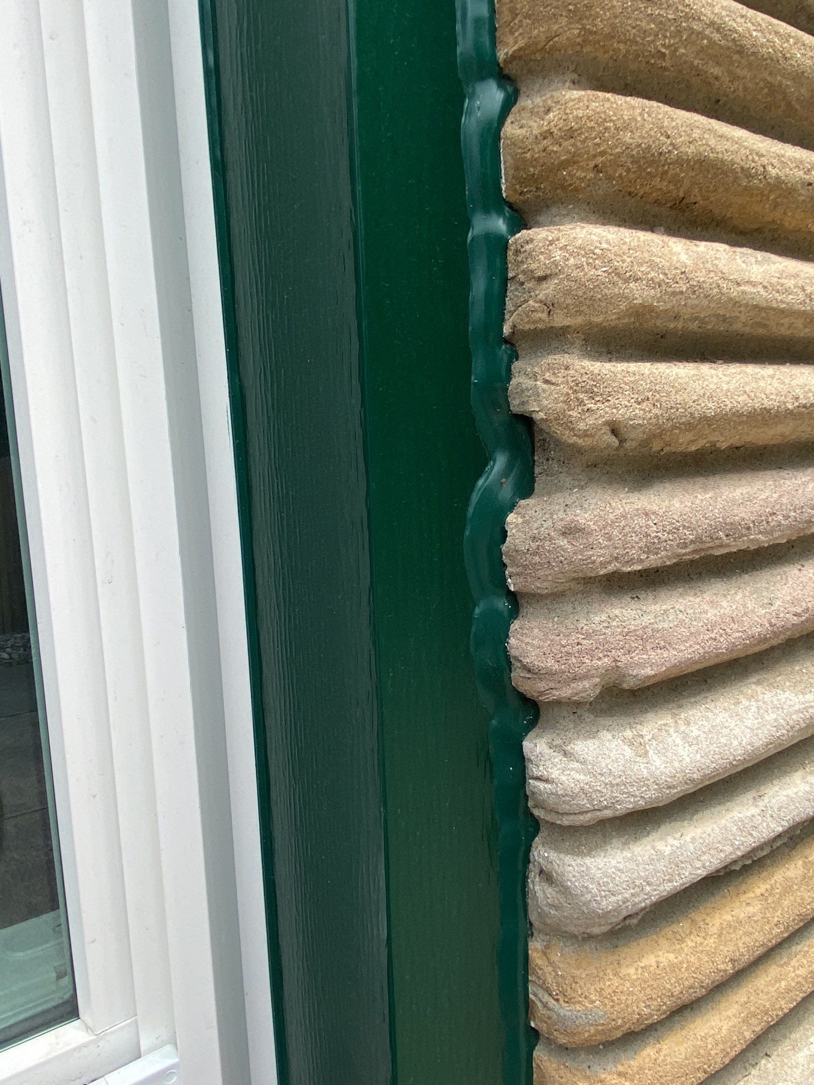 Close-up of a green window frame beside a beige stone wall with vertical blinds inside