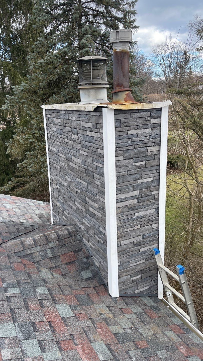 Stone chimney with two metal flues on a shingled rooftop, surrounded by trees.