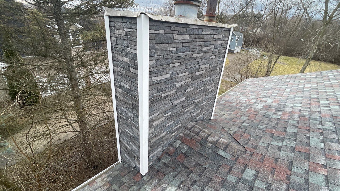 Rooftop chimney with gray stone siding and white trim beside a shingled roof and leafless trees.