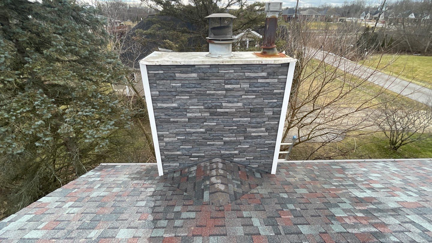 Roof with a gray brick chimney and white trim amid trees on an overcast day