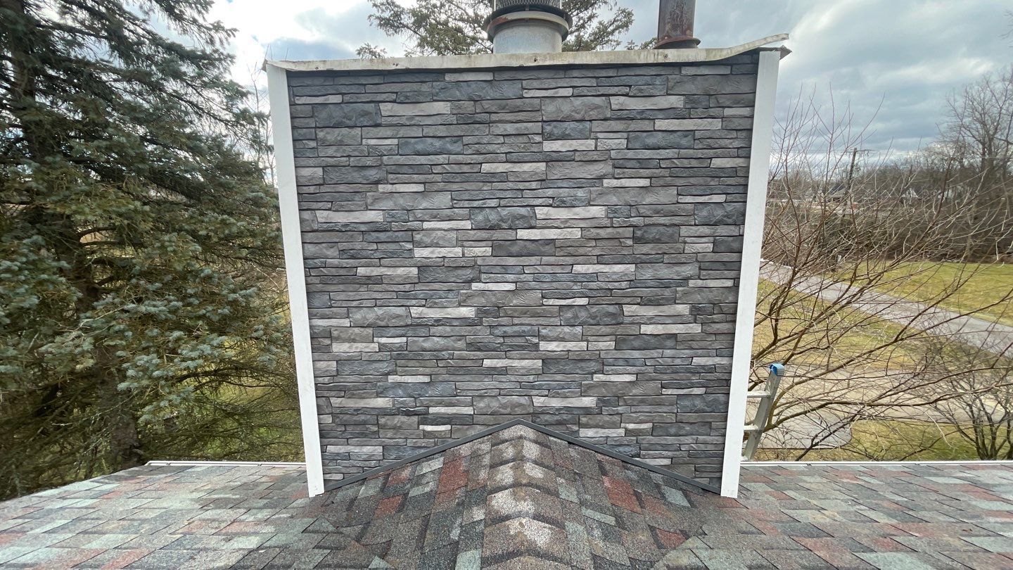 Stone chimney on a roof with gray shingle siding, trees and a road in the background.