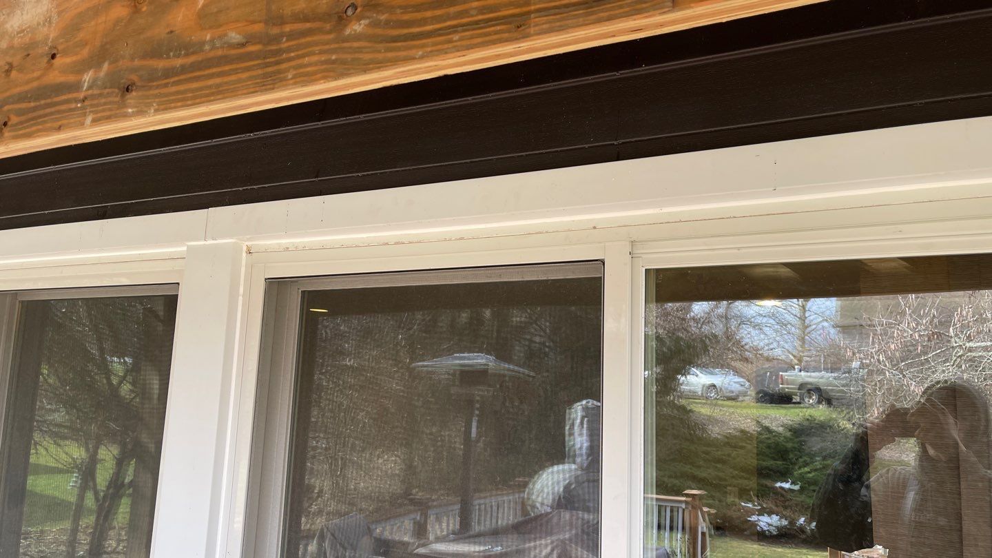 White-trimmed windows under a wooden roof eave, with reflections of trees and a room interior visible.