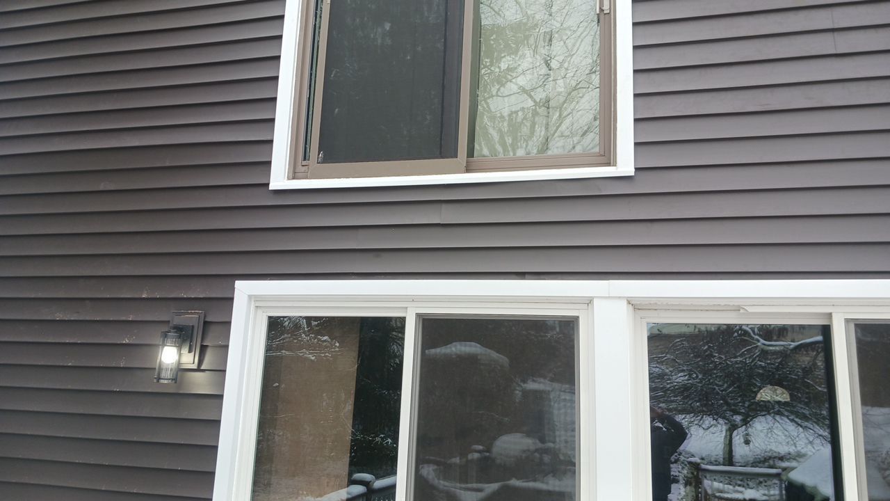 Exterior of a house with dark siding, white-trim windows, a lit wall lantern, and sliding glass doors