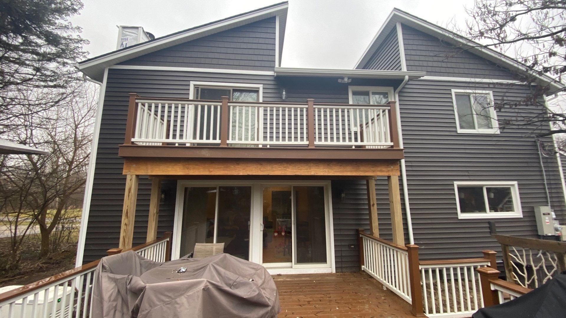 Back view of a dark gray house with a wooden balcony and patio, covered grill, and sliding glass doors