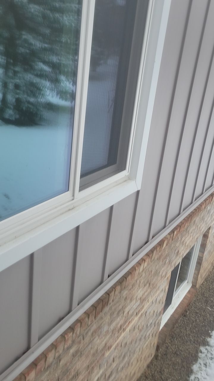 Close-up of a house exterior with a window, gray siding, and a stone foundation in winter.