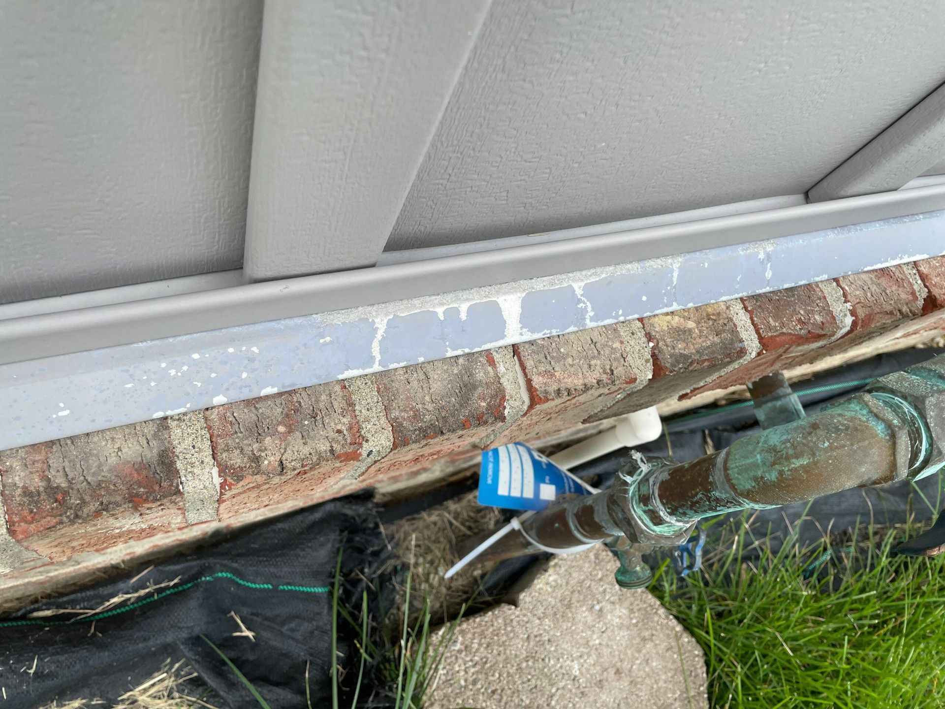 Close-up of a roof edge with gutter, exposed wiring, and a broken pipe beside grass and brick wall