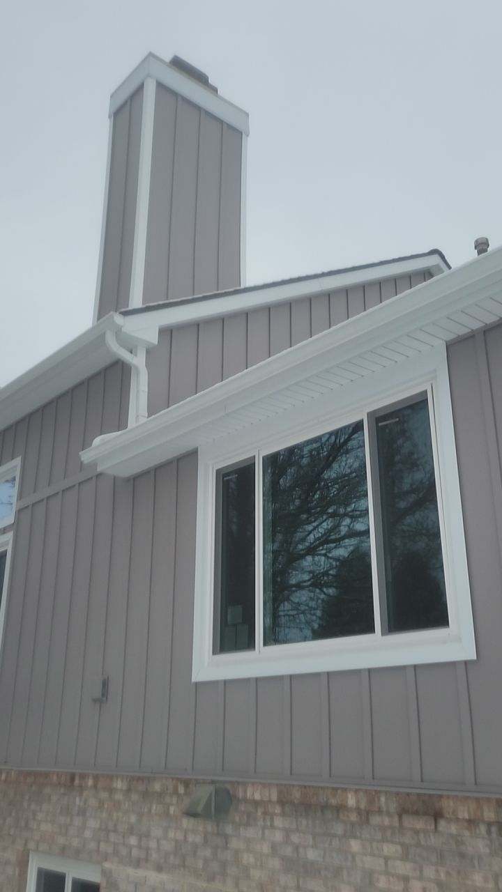 Tan-sided house exterior with white trim, a large window, and a brick foundation under an overcast sky