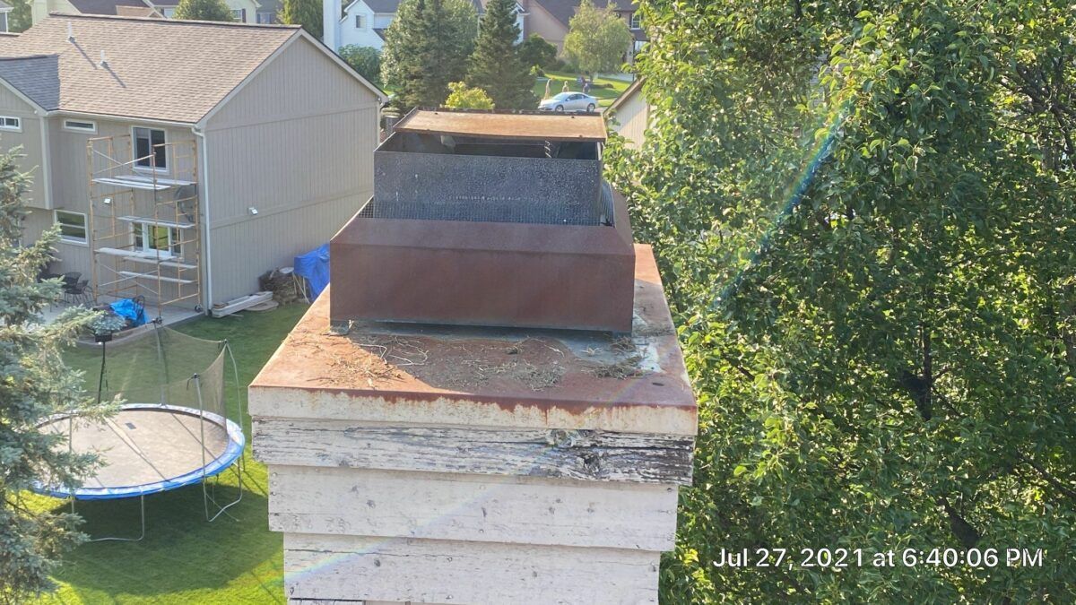 Two stacked chimney caps on a white rooftop with leafy trees and neighboring houses in the background