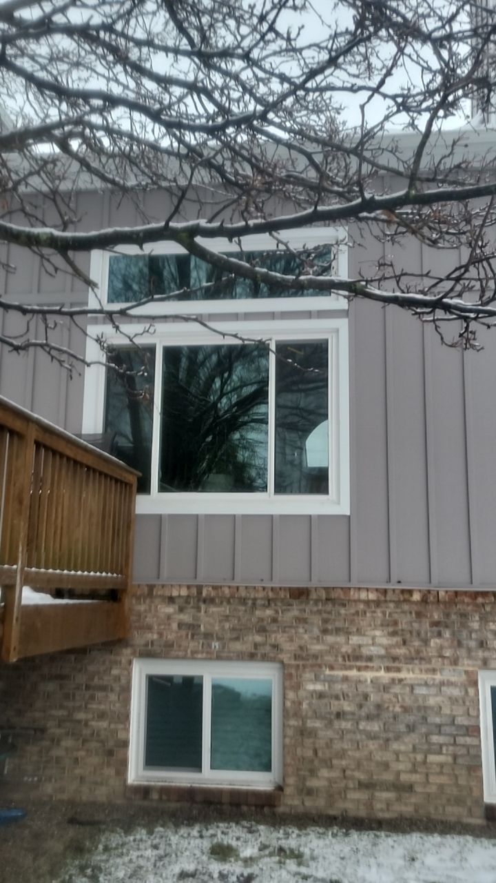 Two-story house exterior with gray siding, brick base, white windows, and bare tree branches overhead