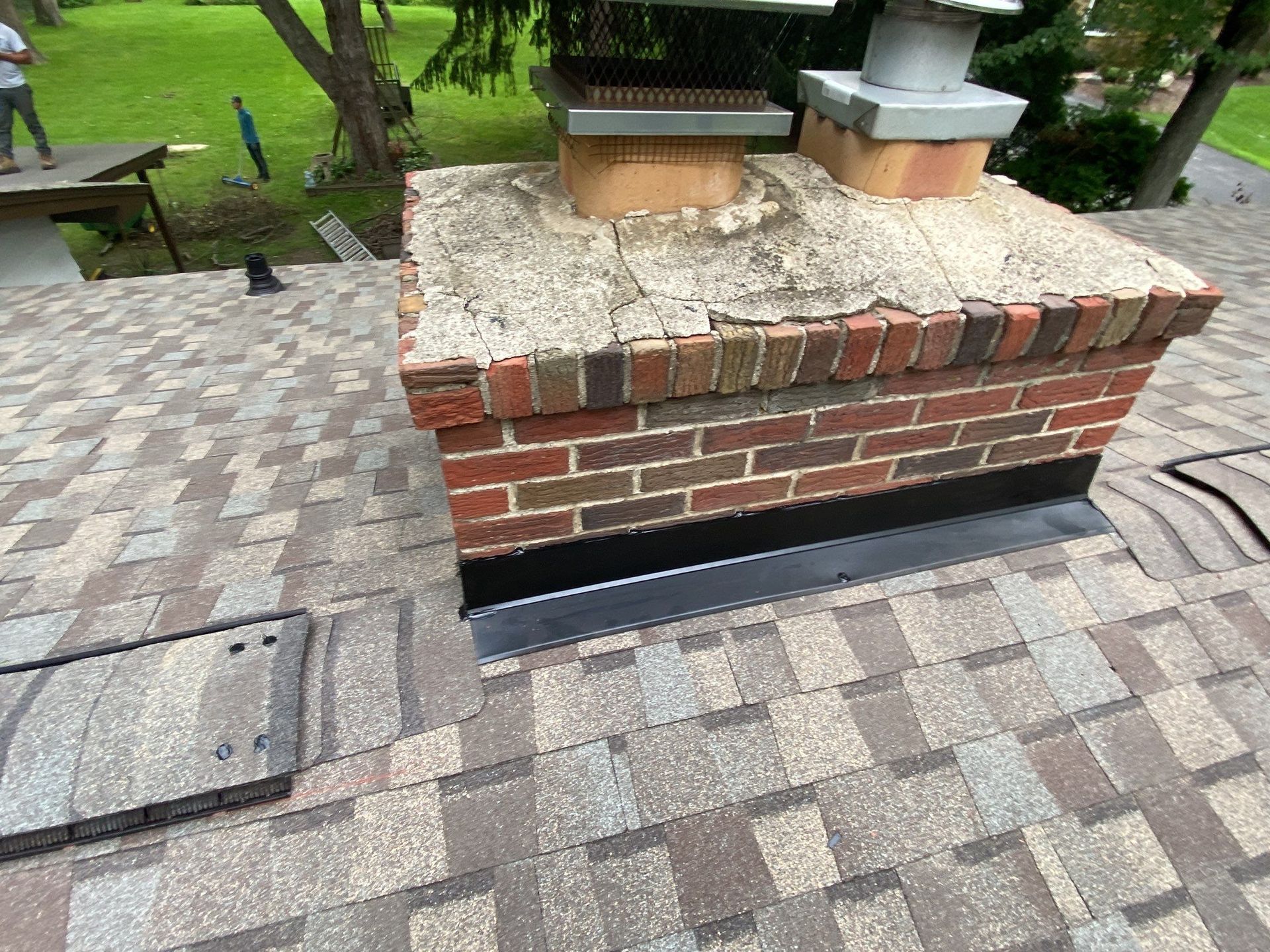 Brick outdoor grill with metal chimney on a cobblestone patio near grass and trees