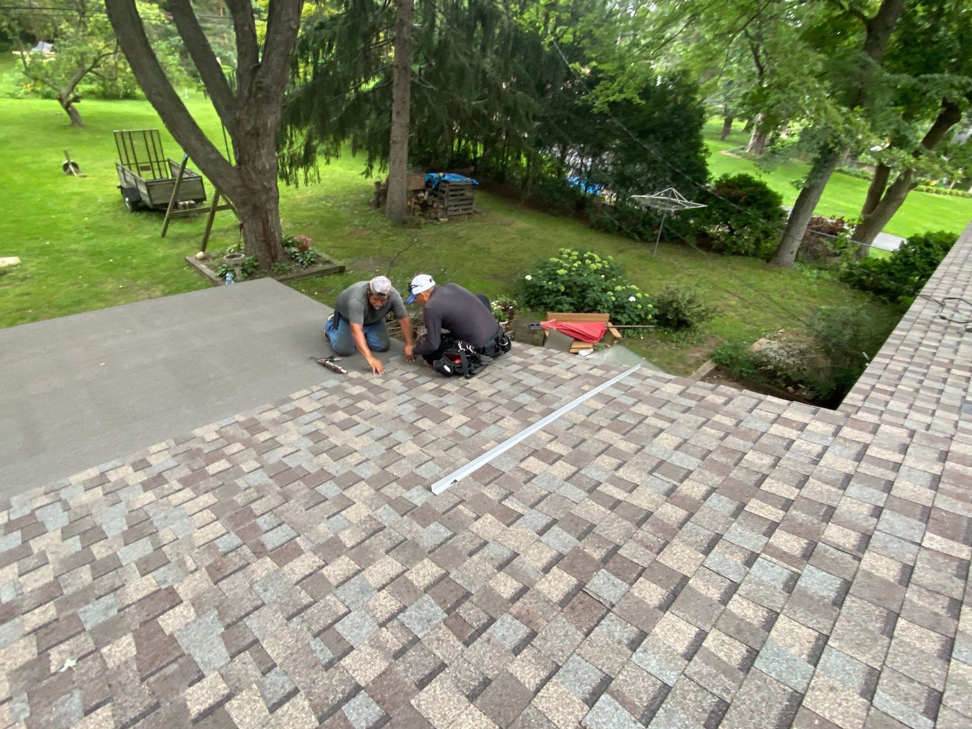 Two workers crouch on a brick patio beside a green backyard with trees and shrubs.