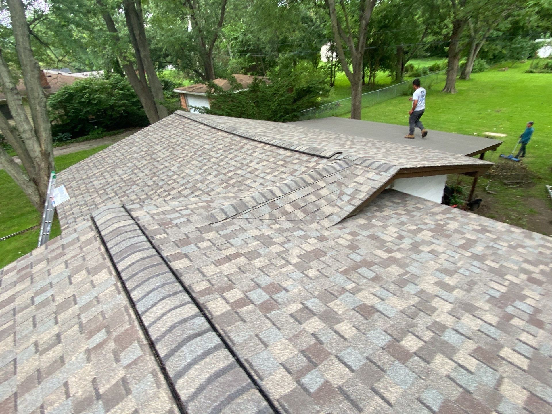Shingled roof in a wooded park with two workers nearby and a ladder on the roof