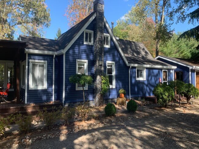 Blue house with white trim, front porch, and potted plants under tall trees in daylight