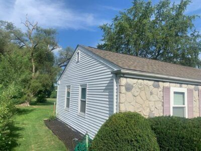 Small house with gray siding and stone exterior, beside a green lawn and trees under a blue sky