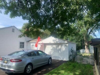 Silver car parked in a driveway beside a white garage and house, with a red flag and tree overhead