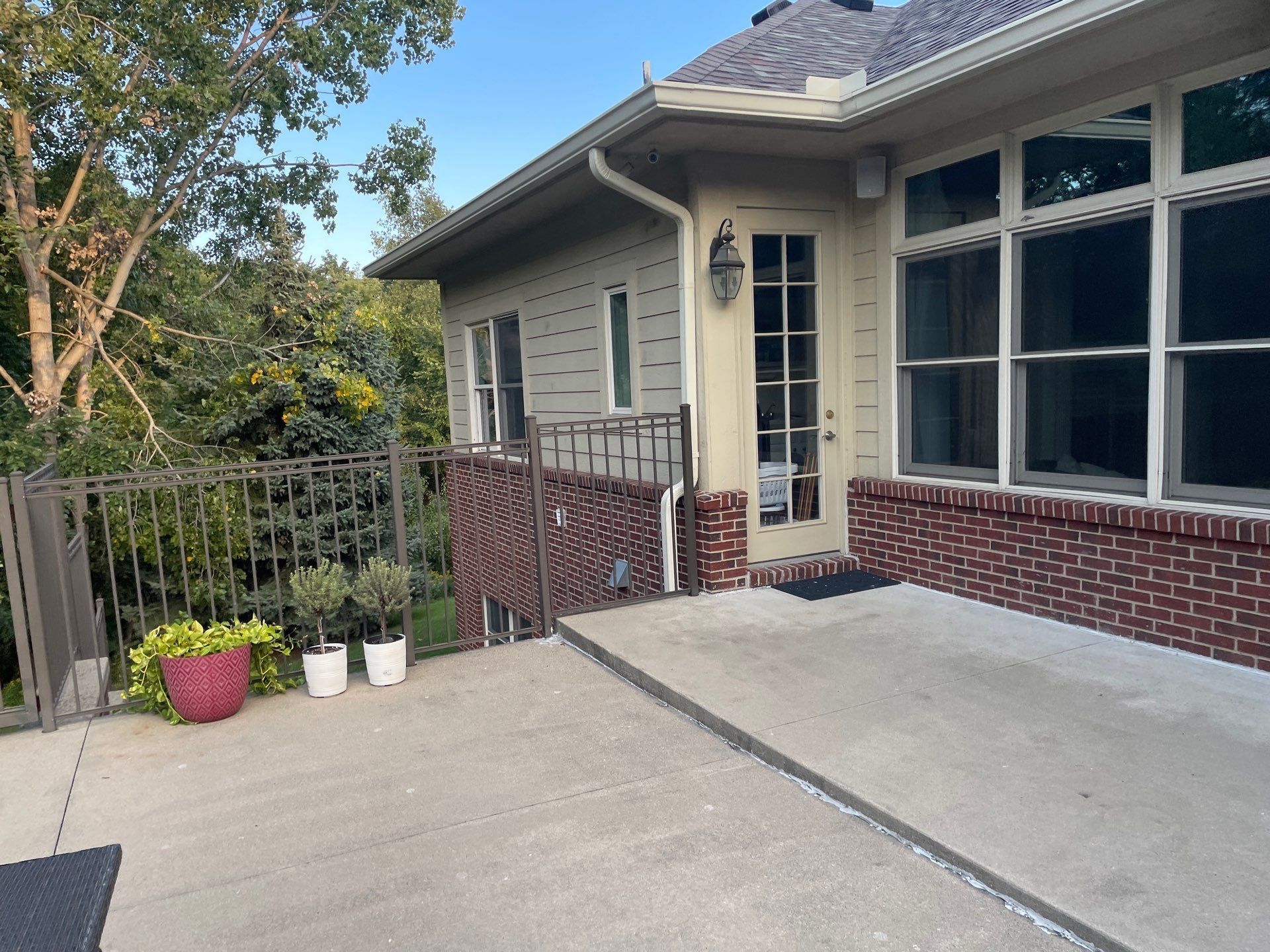 Patio beside a house with railing, potted plants, and large windows on a sunny day