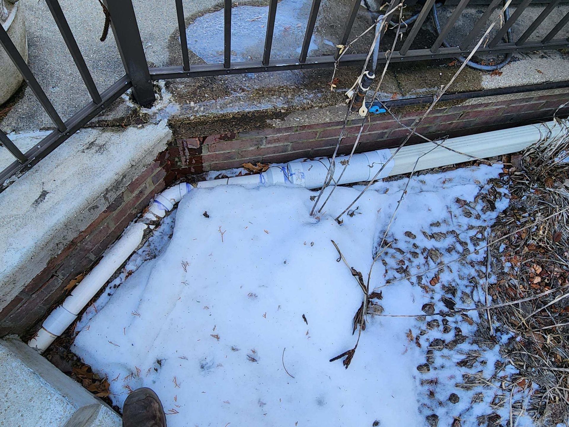 Snow-covered patio beside metal railing and a brick wall with scattered leaves.
