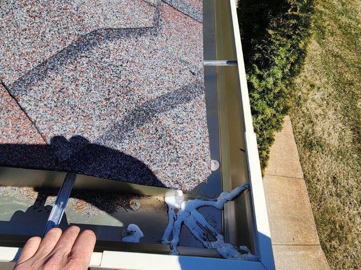 Hands working on a shingled roof near the gutter, with a ladder and yard visible below.