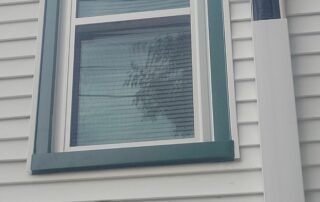 White house exterior with a small window framed in teal trim and blinds inside