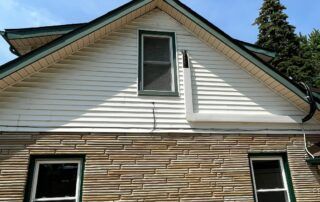 Side of a house with white siding, tan stone facade, and two windows beneath a peaked roof.
