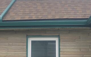 House exterior with tan siding, brown shingle roof, and a white-framed window with blinds