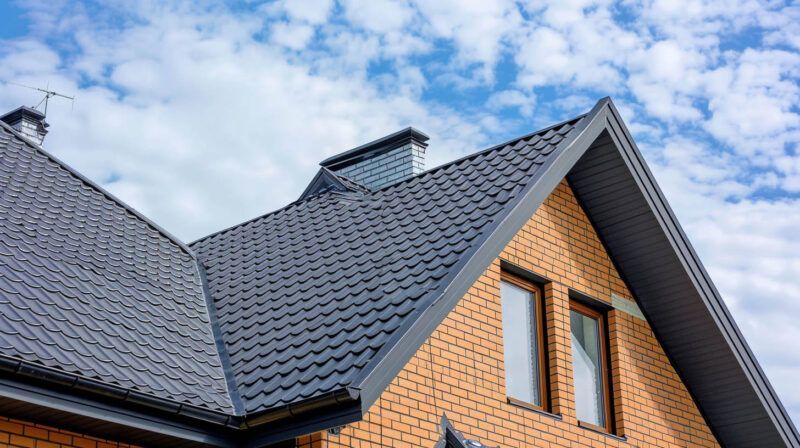 Close-up of a house roof with dark gray shingles and brick gables under a partly cloudy sky