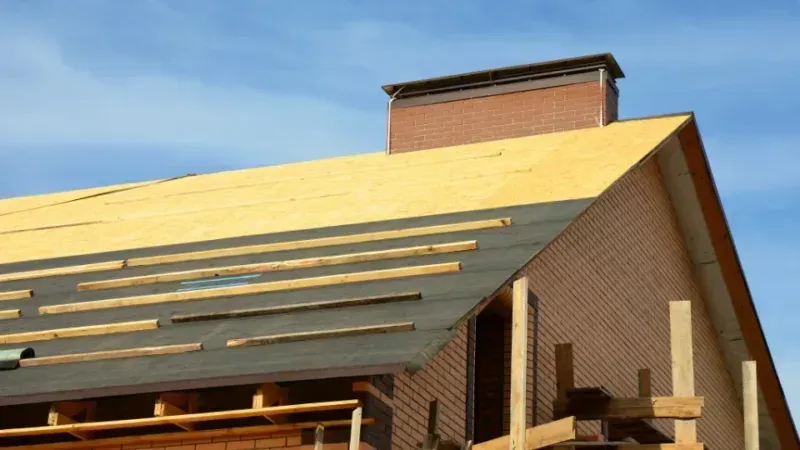 Brick building roof under construction with yellow sheathing and a chimney against a blue sky