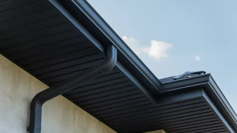 House eave with dark soffit and gutter against a light wall and blue sky