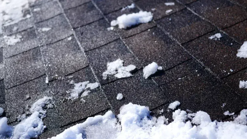 Snow piled on a dark shingled roof, with scattered patches of snow across the surface.