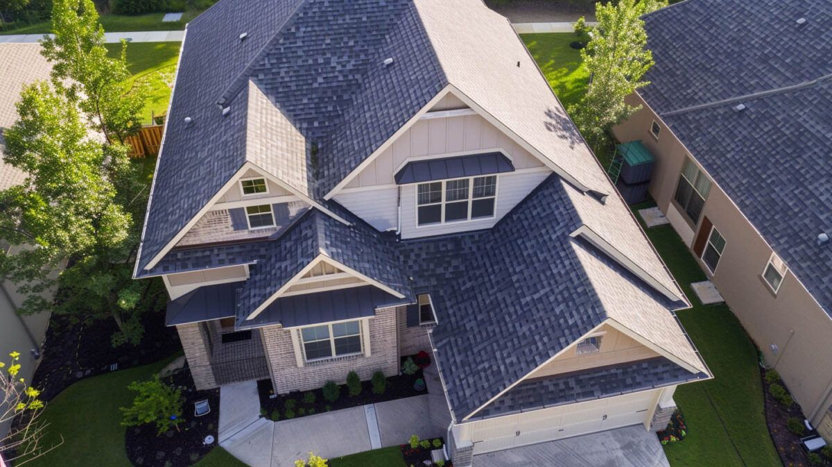 Aerial view of a gray-roofed suburban house with dormer windows and a front porch surrounded by trees.