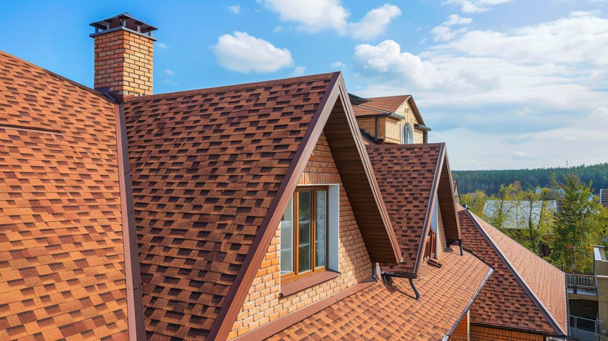 Red-tiled rooftops with dormer windows and a chimney against a blue sky and distant trees