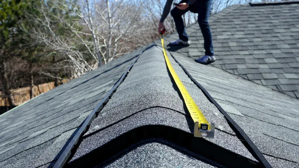 Worker measuring a roof ridge with a yellow tape measure on gray shingles