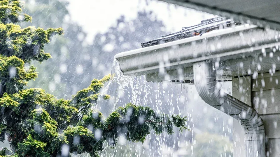 Rain pouring off a white house roof beside green tree branches.