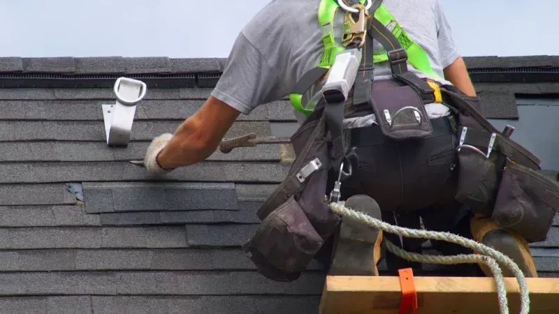 Worker on a roof installing dark shingles, wearing a safety harness and tool belt.