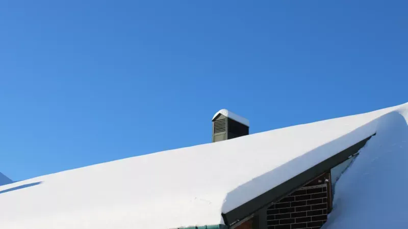 Snow-covered roof with a black chimney against a clear blue sky