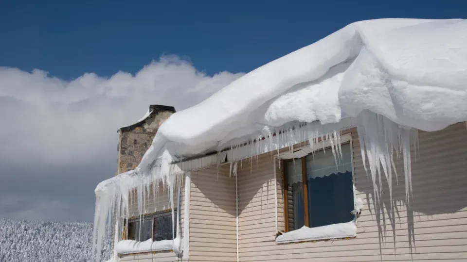 Snow-covered house roof with long icicles in a winter landscape under a cloudy sky