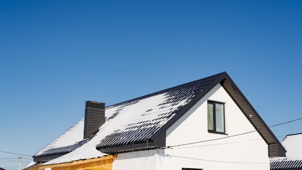 Snow-covered dark roof of a white house under a clear blue sky